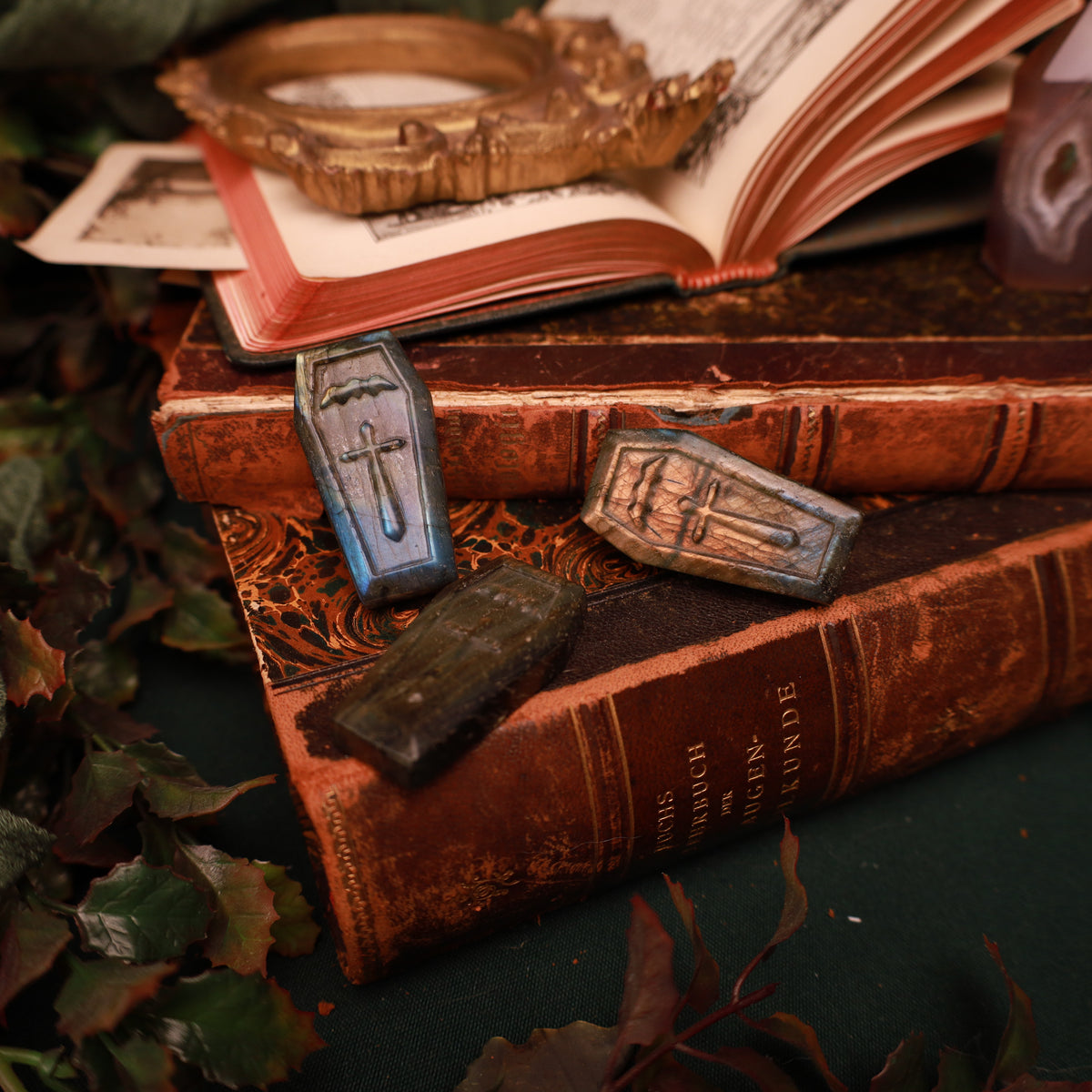 Labradorit-Sarg mit eingraviertem Kreuz und kleiner Fledermaus, kupfergoldenen und blauen Reflexen, aufrecht vor alten Büchern arrangiert.