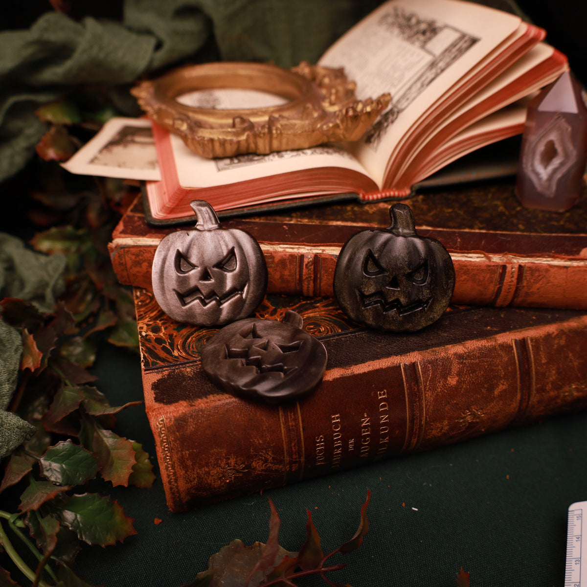 Drei dunkle Obsidian-Halloweenkürbisse mit schrägen Augen und gezackten Mündern auf alten Büchern, zwei aufrecht und einer liegend.
