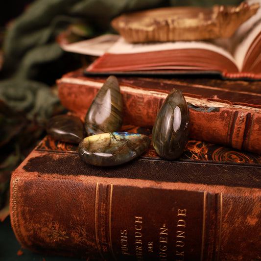 Vier glänzende Labradorit-Steine in Tropfenform auf alten Büchern, zwei aufrecht und zwei liegend, mit goldfarbenen, grünlichen und blauen Reflexen.