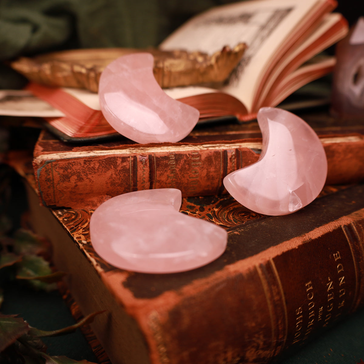 Drei rosa Rosenquarz-Mondsicheln mit glänzender Oberfläche und hellen inneren Strukturen auf alten Büchern, zwei aufrecht und eine liegend.