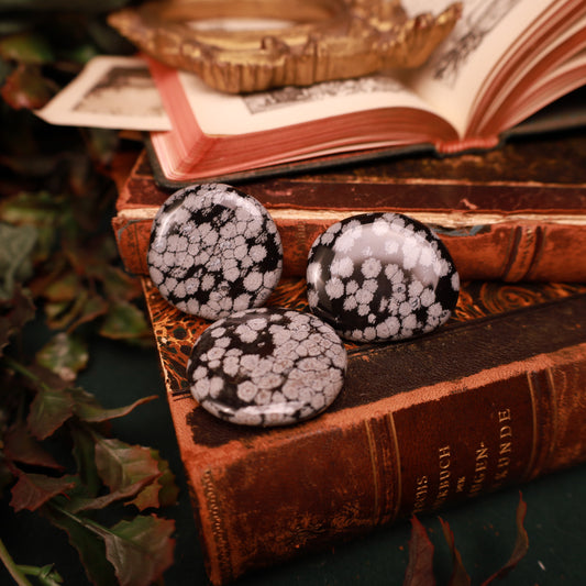 Drei glänzende Handschmeichler aus Schneeflockenobsidian auf einem Bücherstapel. Die schwarzen, runden Steine zeigen unterschiedlich dicht verteilte helle Muster.
