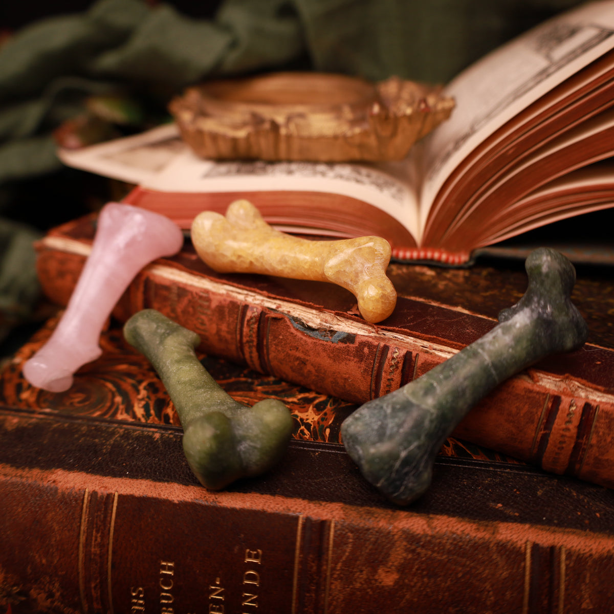 Vier glänzende Steinschnitzereien in Knochenform in Rosa, Gelb und zwei Grüntönen, auf alten Büchern arrangiert.