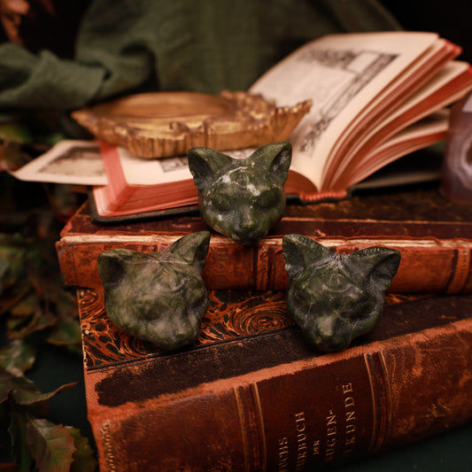 Drei dunkelgrüne Katzenköpfe aus Jade mit spitzen Ohren und einem zusätzlichen Auge auf der Stirn, auf alten Büchern arrangiert.