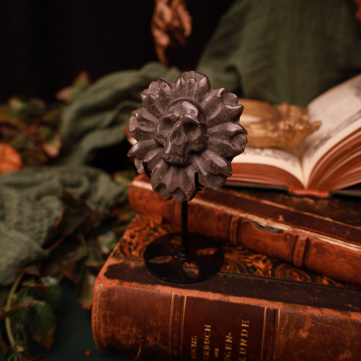 Deathflower aus Silberobsidian mit zentralem Totenkopf und plastischen Blütenblättern, auf einem schwarzen Metallständer vor alten Büchern präsentiert.