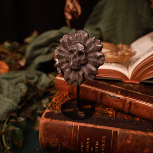 Deathflower aus Silberobsidian mit zentralem Totenkopf und plastischen Blütenblättern, auf einem schwarzen Metallständer vor alten Büchern präsentiert.